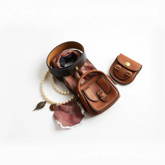 Vintage-inspired accessories including a leather belt, scarf, necklace, and brooches on a neutral tabletop.